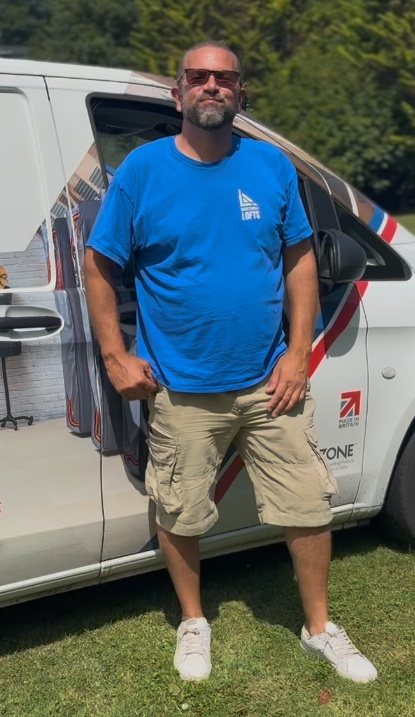 Man wearing sunglasses, a bright blue T-shirt with white logo, beige cargo shorts, and white sneakers standing on grass beside a white vehicle with red and blue graphics.