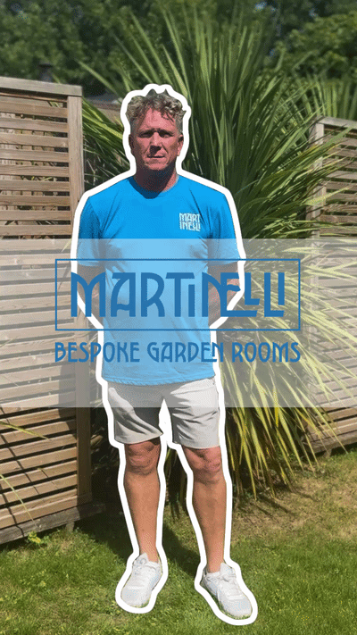Man standing on grass in front of wooden garden fencing and green foliage wearing a blue T-shirt, light shorts, and white sneakers with a white outline around him and a semi-transparent overlay with the text 'Martinelli Bespoke Garden Rooms'.