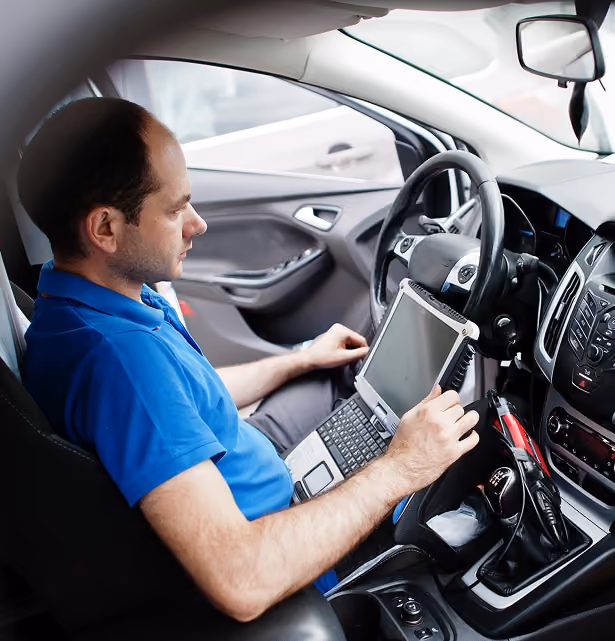 Man sitting in a car driver's seat using a laptop for vehicle diagnostics or programming.