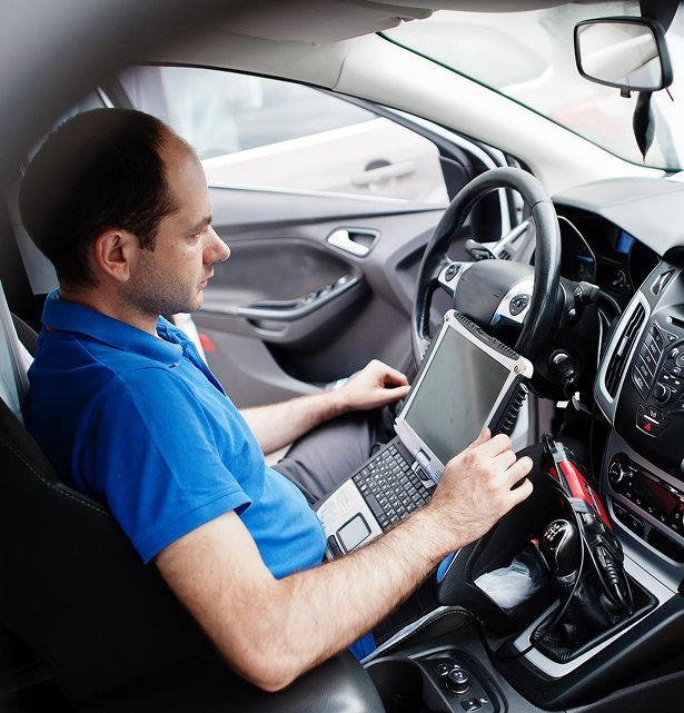 Man sitting in a car driver's seat using a laptop for vehicle diagnostics or programming.