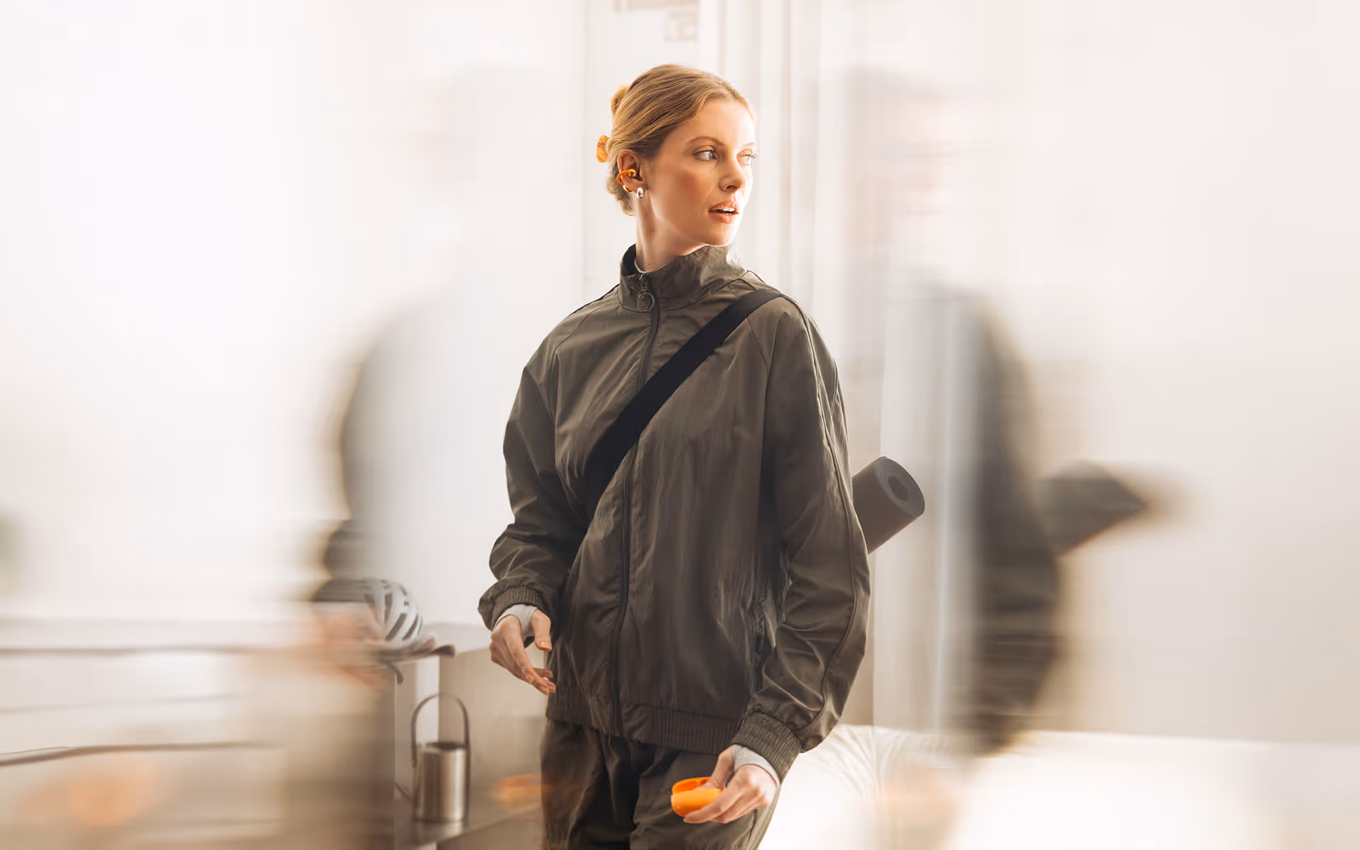 Woman in a dark jacket with earplugs and yoga mat looks to the side indoors with blurred foreground figures.