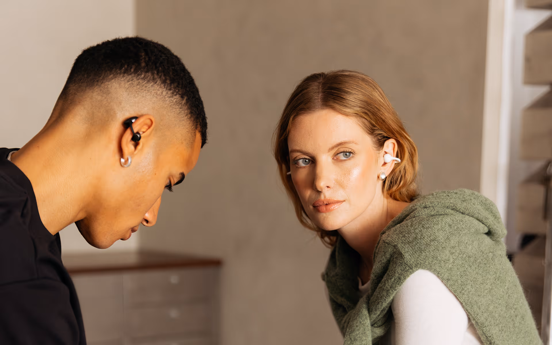 Man and woman wearing wireless earbuds in a neutral indoor setting, the woman looks at the camera.