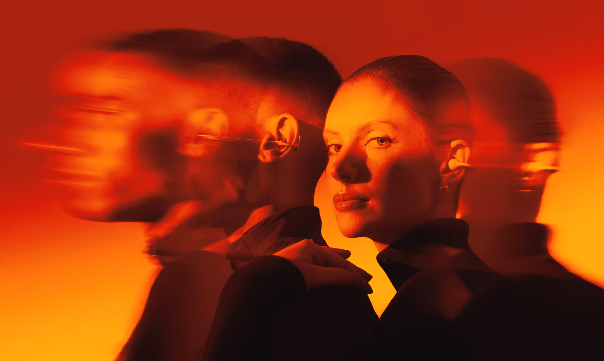 Portrait of a woman facing forward with blurred side profile of a man in warm red-orange lighting.