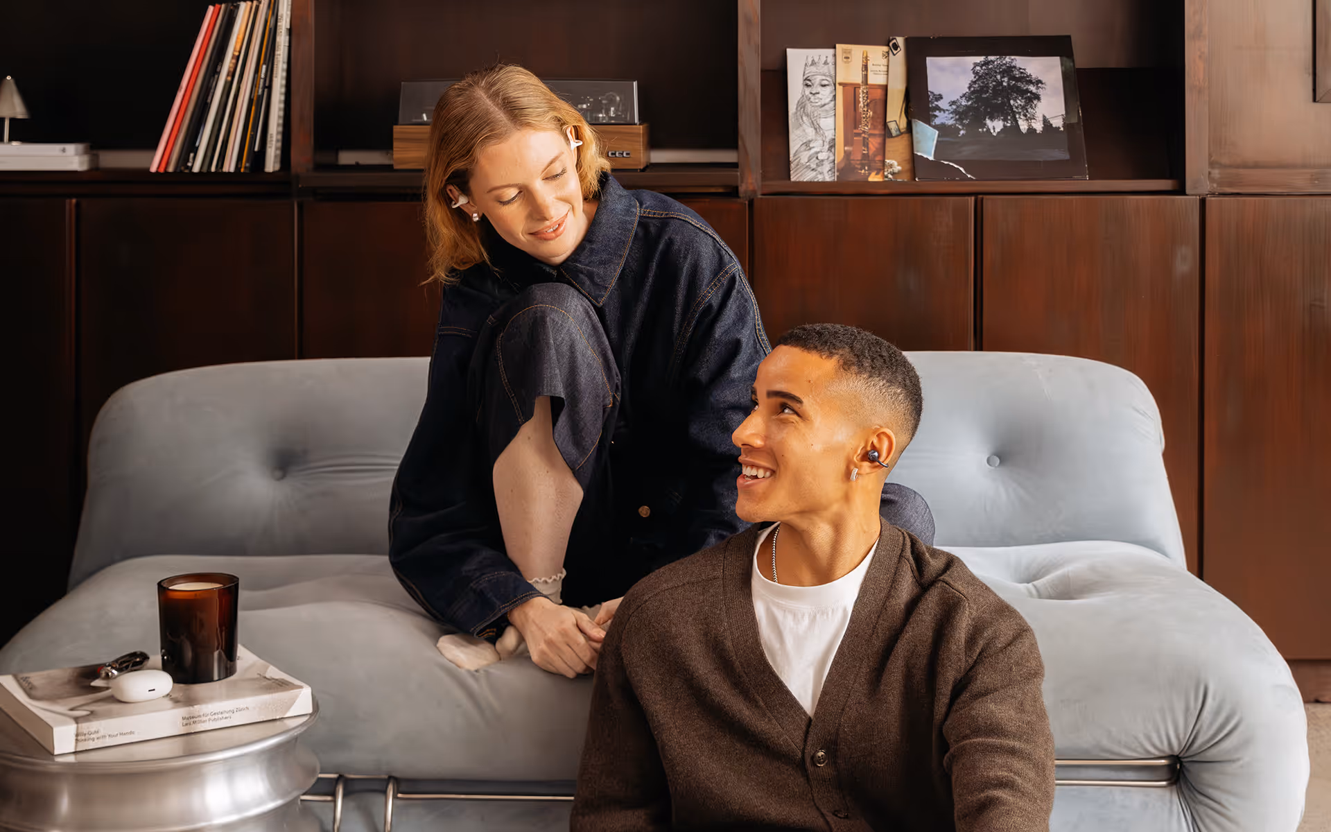 Smiling young woman and man wearing wireless earbuds sitting on a light gray couch in a cozy room.