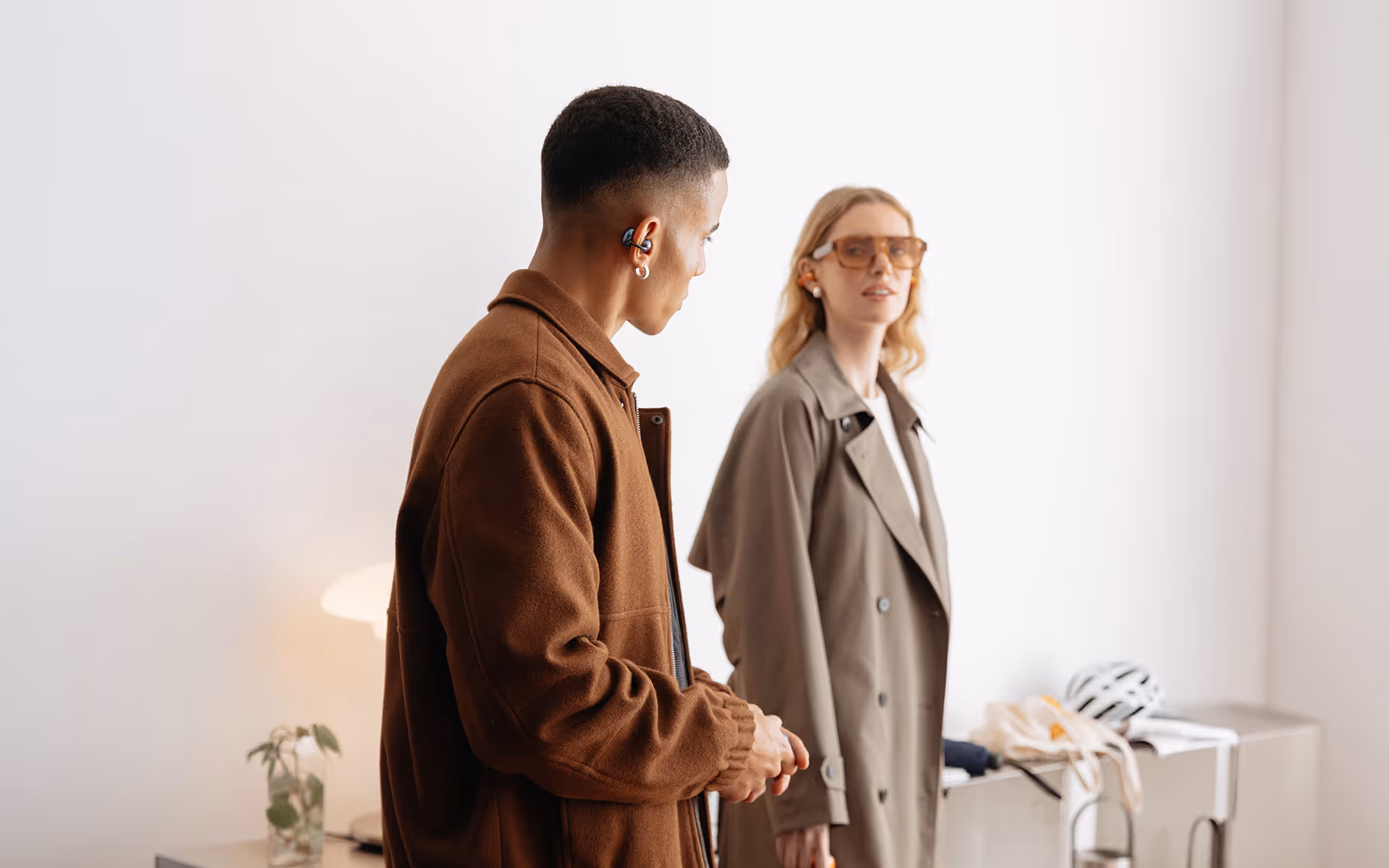 Man in brown jacket listens with earbuds, looks at woman wearing glasses and trench coat indoors.