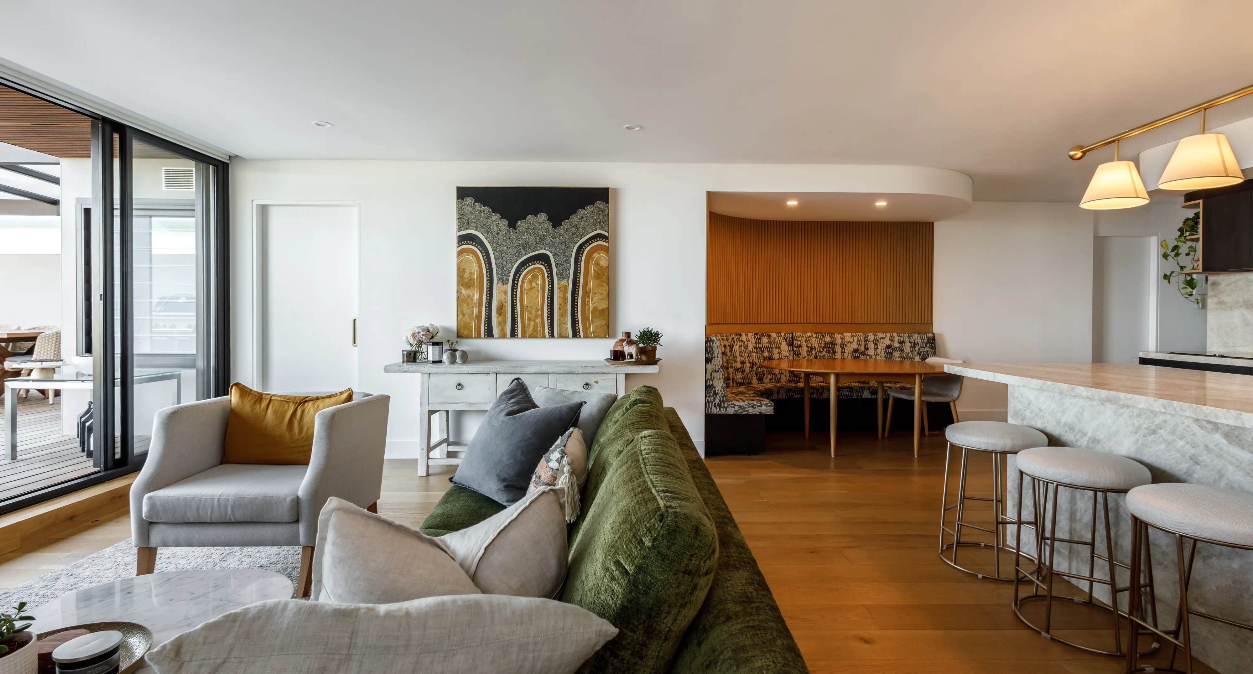 Modern living room with green sofa, beige armchair, dining nook, and kitchen island with bar stools.