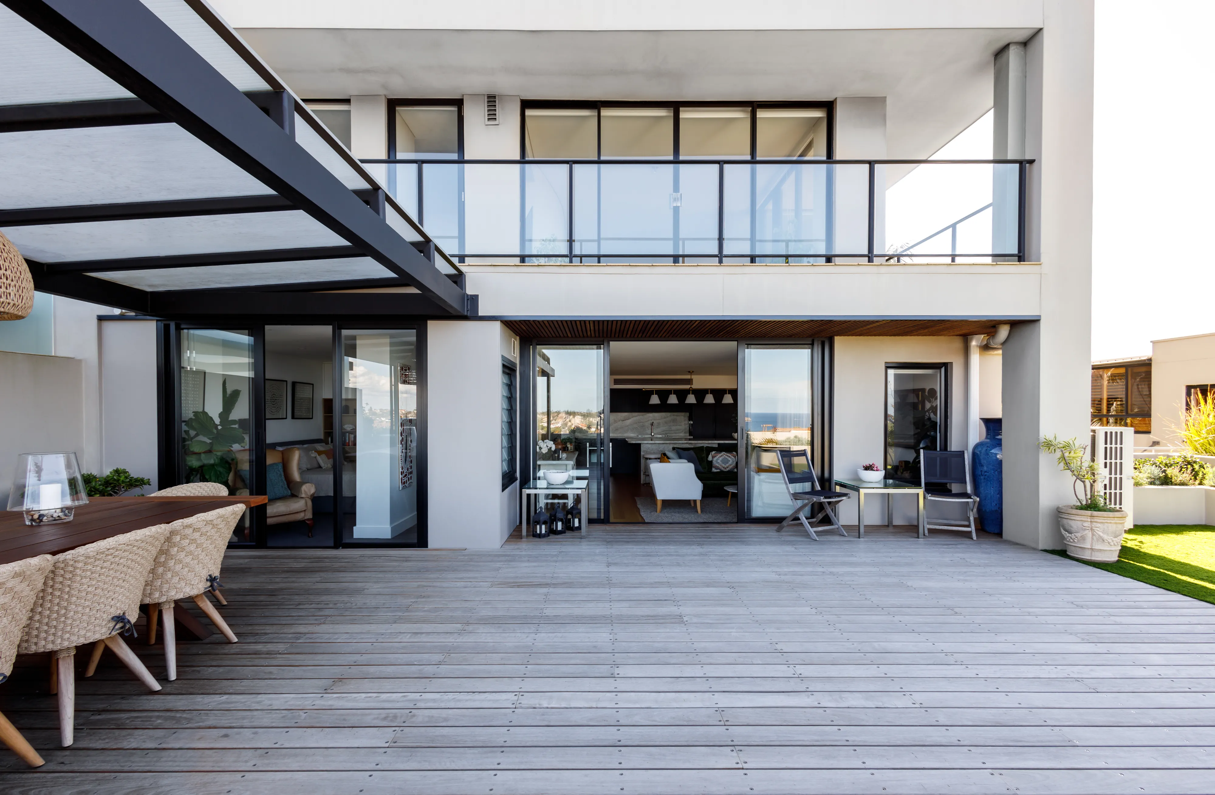 Modern two-story patio with wooden deck, outdoor dining table, and glass railing on upper balcony.