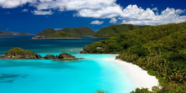 Turquoise ocean water meets white sandy beach surrounded by lush green hills under a cloudy blue sky.
