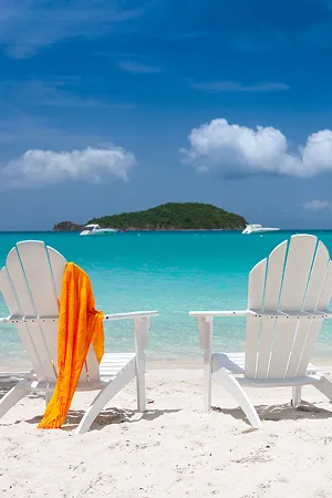 Two white beach chairs on sand with an orange towel, turquoise sea, island, and blue sky with clouds.