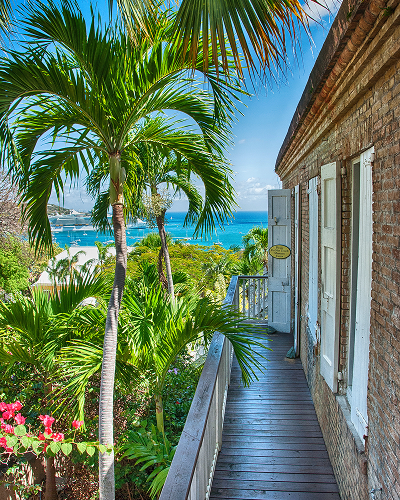 Balcony with wooden floor by brick wall overlooking palm trees and a blue ocean under a clear sky.