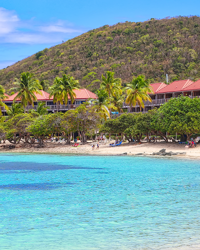 Clear turquoise water, beach with palm trees, red-roofed buildings, and green hillside backdrop.