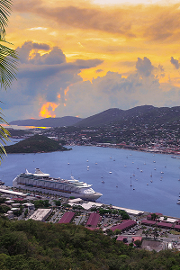 Sunset view of a harbor with cruise ship, boats, hills, and scattered buildings under cloudy sky.