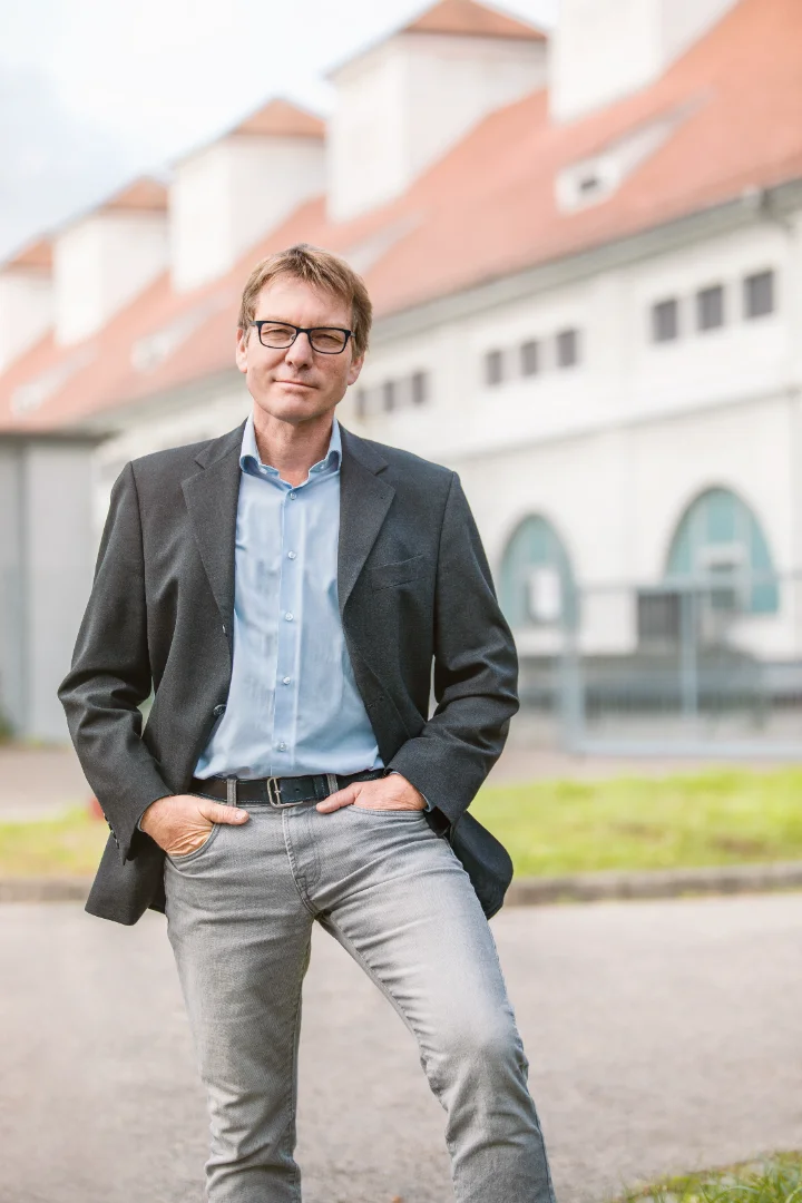 Mann mit Brille trägt Sakko und Hemd, lehnt mit Händen in den Taschen vor einem Gebäude.