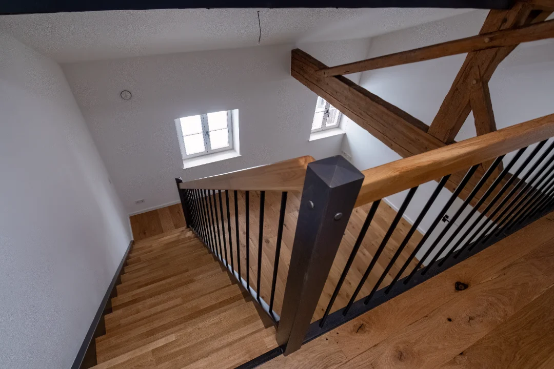 Holztreppe mit schwarzem Geländer und sichtbaren Holzbalken in einem weißen Raum mit Fenstern.