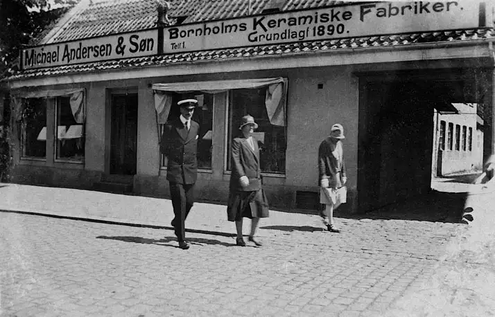 Sort-hvid foto af tre personer, der går foran en bygning med skilt for Bornholms Keramiske Fabriker.