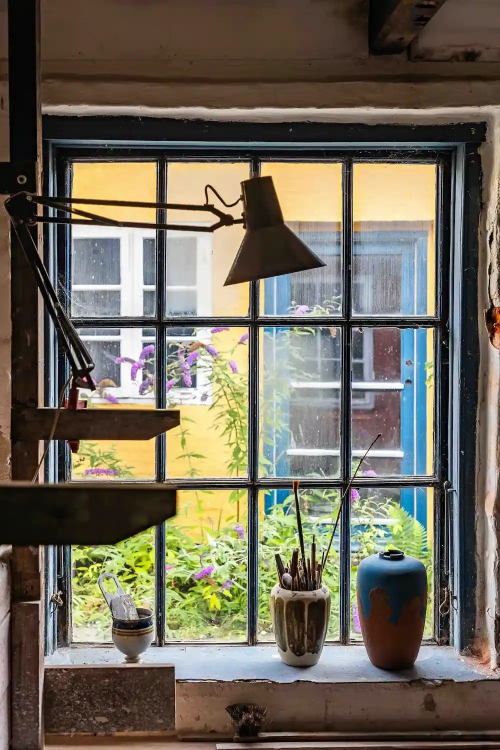 Vindu med blomsterpotter og skrivebordslampe med gule og blå huse udenfor.