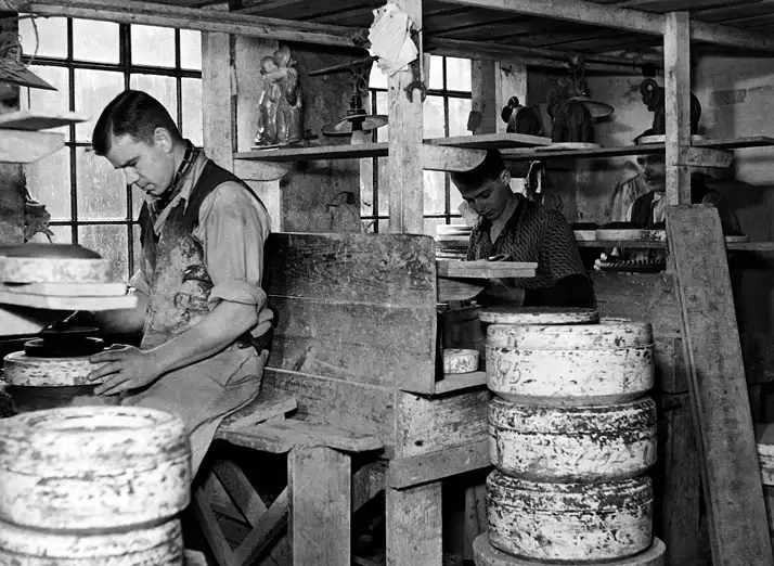 To mænd arbejder i en keramisk fabrik med drejeskiver og lerfigurer i et gammelt værksted.