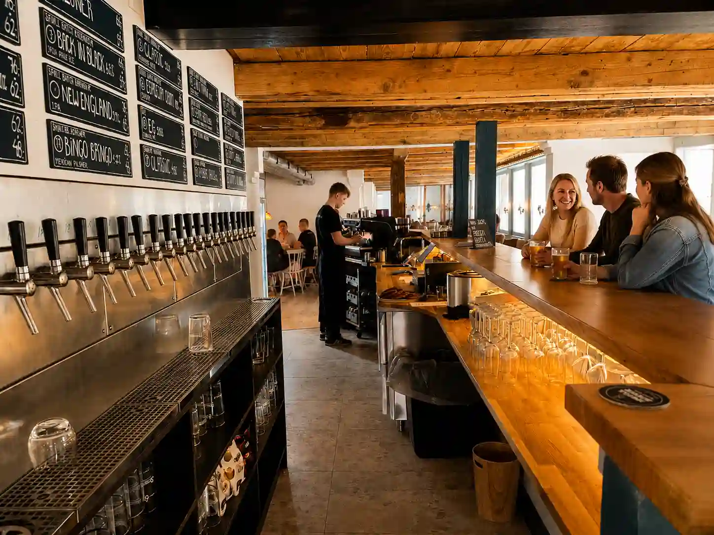 Tre mennesker sidder ved en træbar i en hyggelig bar med fadølsanlæg og en bartender.