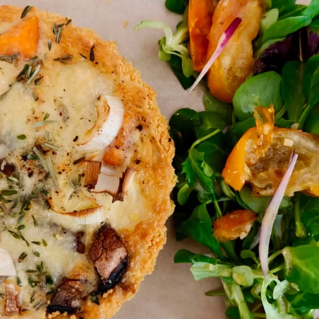 Quiche med ost, løg og svampe ved siden af en salat med grønne blade og bagte grøntsager.