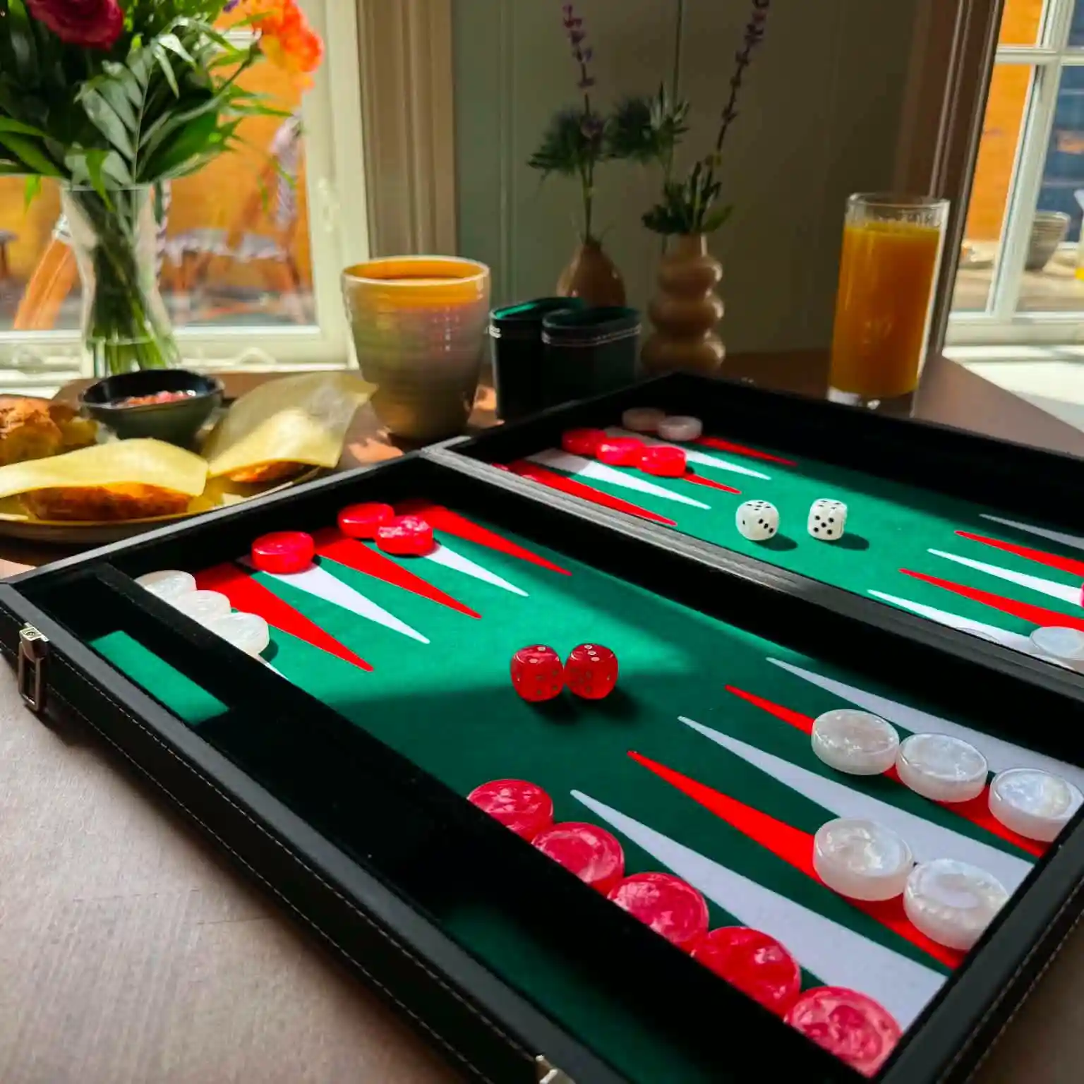 Backgammonspil med røde og hvide brikker på et bord med drikkevarer og blomster i baggrunden.