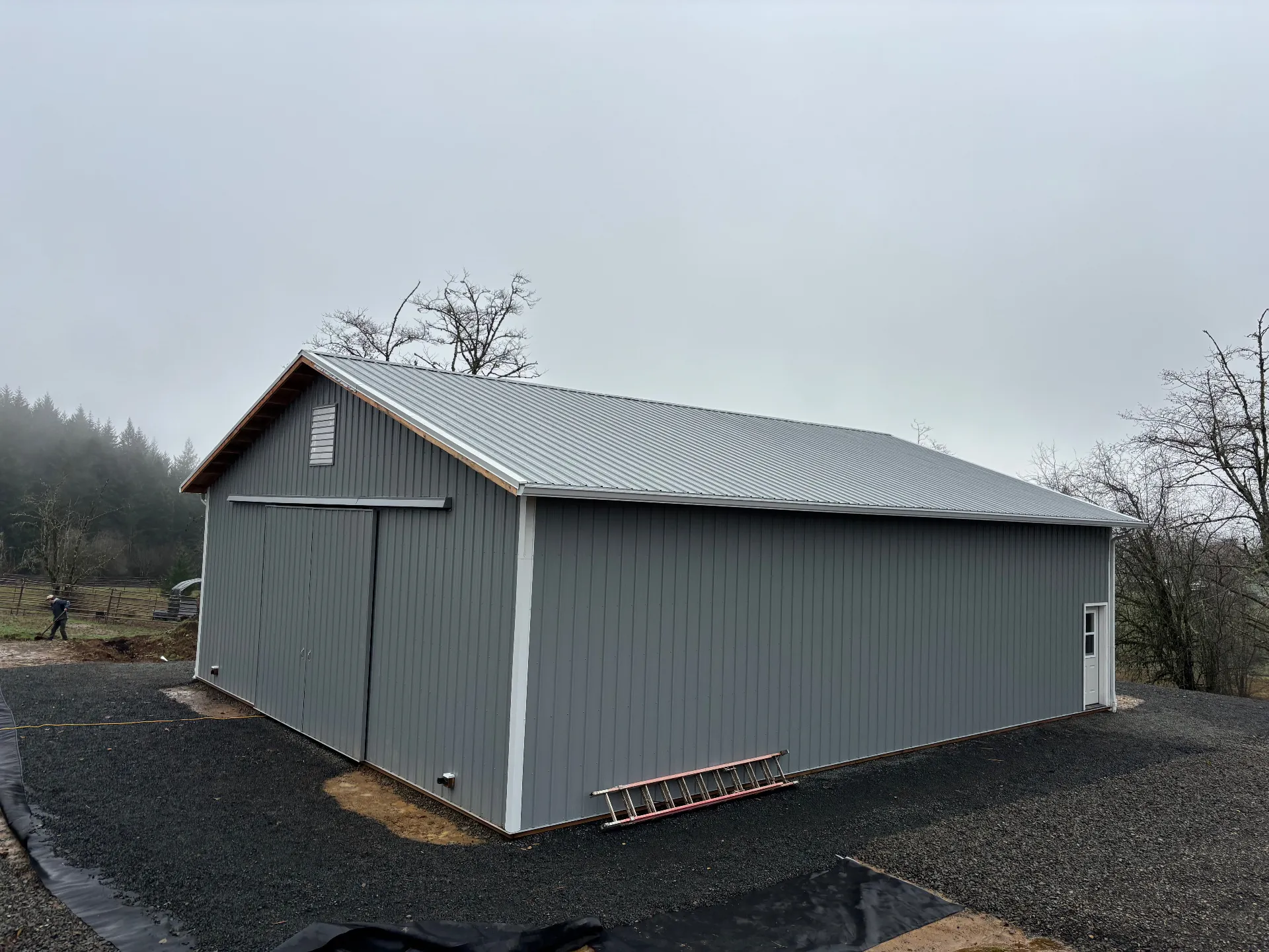 Large gray metal barn with sliding doors and a side door on a gravel base under a cloudy sky.