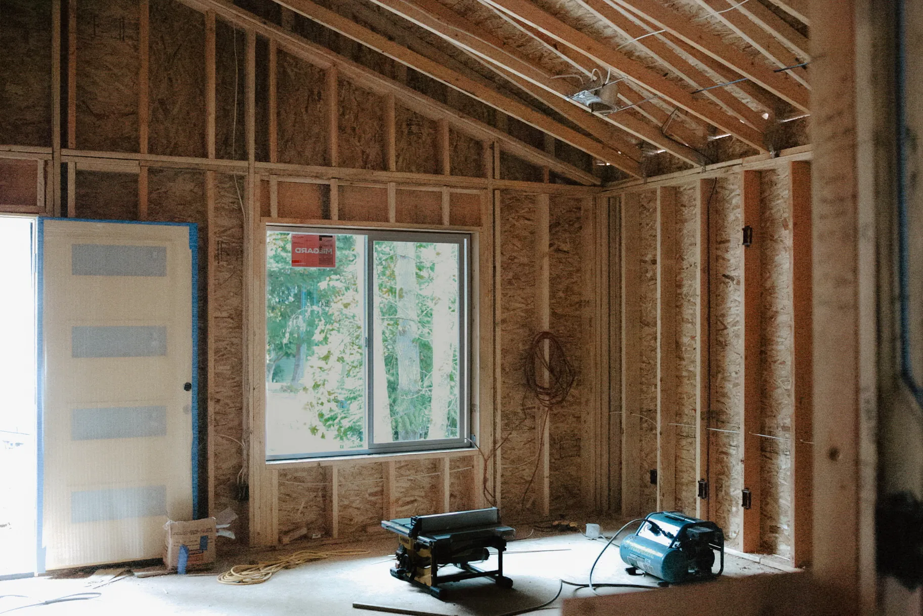 Interior view of a room under construction with exposed wooden framing, a window, and construction tools on the floor.