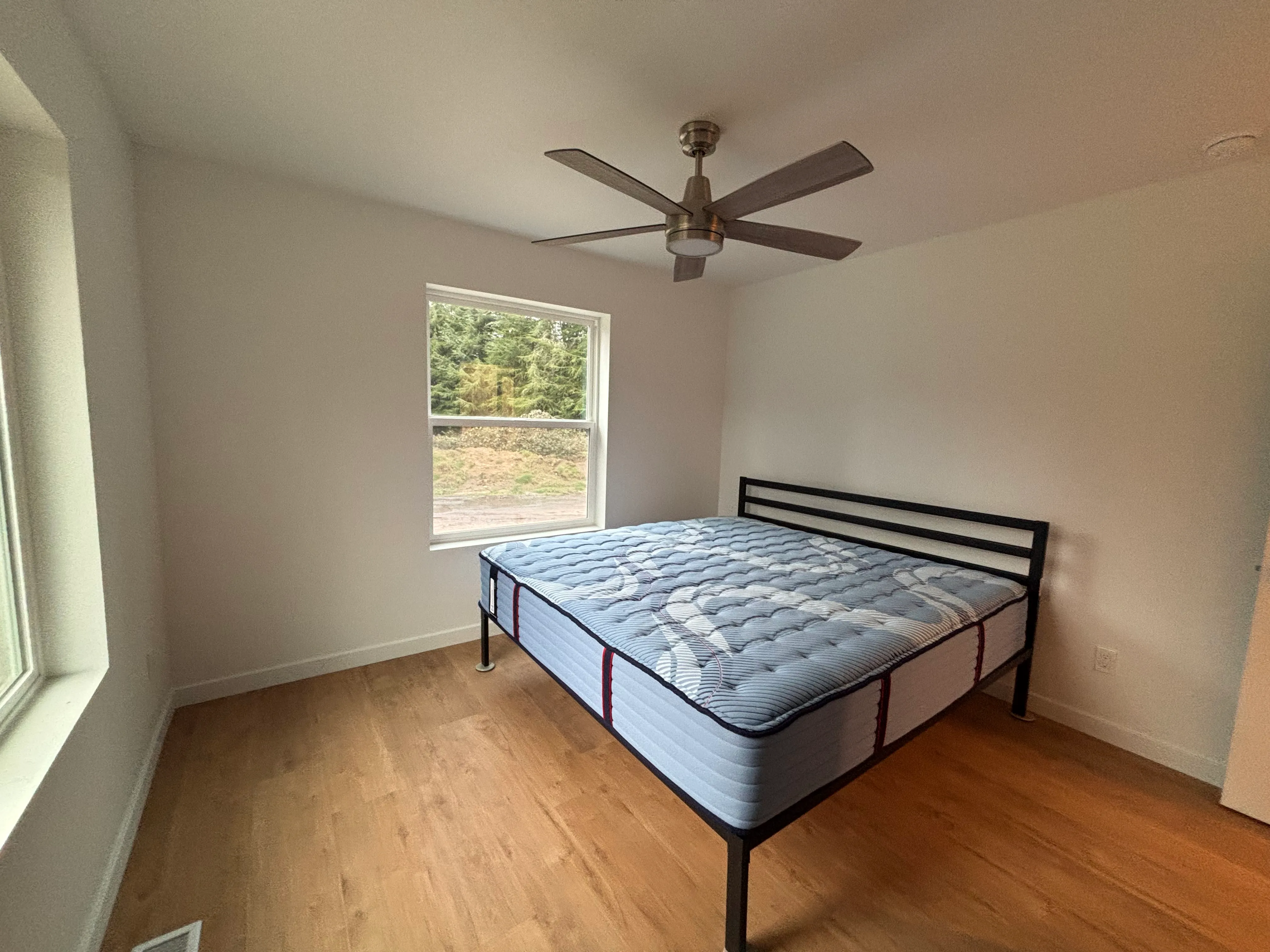 Minimalist bedroom with a mattress on a black metal frame, wood floor, ceiling fan, and window showing outdoor greenery.