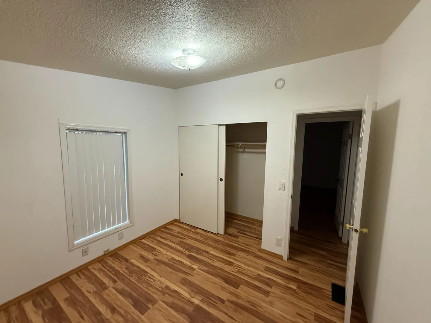 Empty room with wood flooring, white walls, a window with vertical blinds, a closet with sliding doors, and an open door.