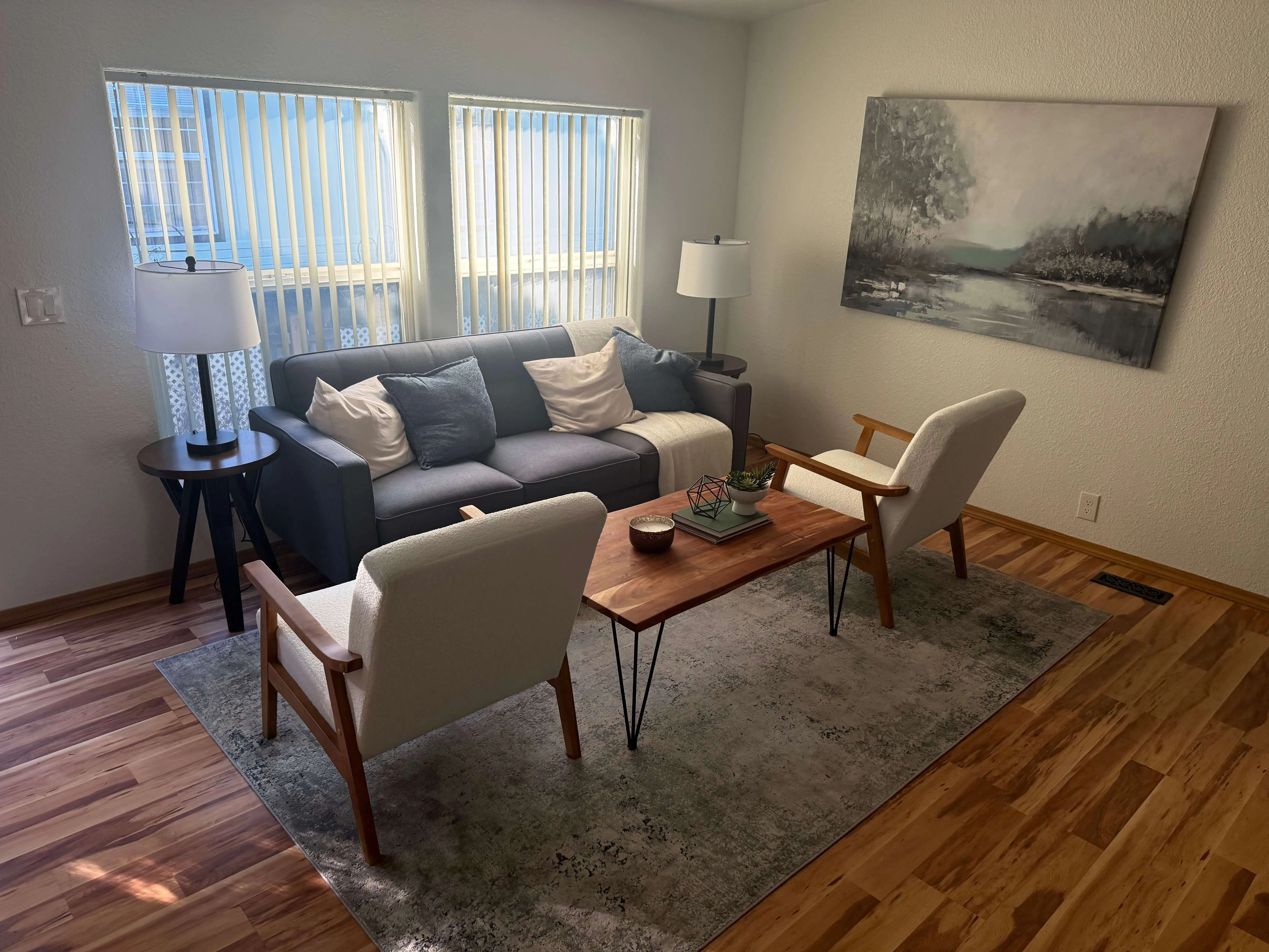 Living room with gray sofa, two cream armchairs, wooden coffee table, lamps, and landscape painting.