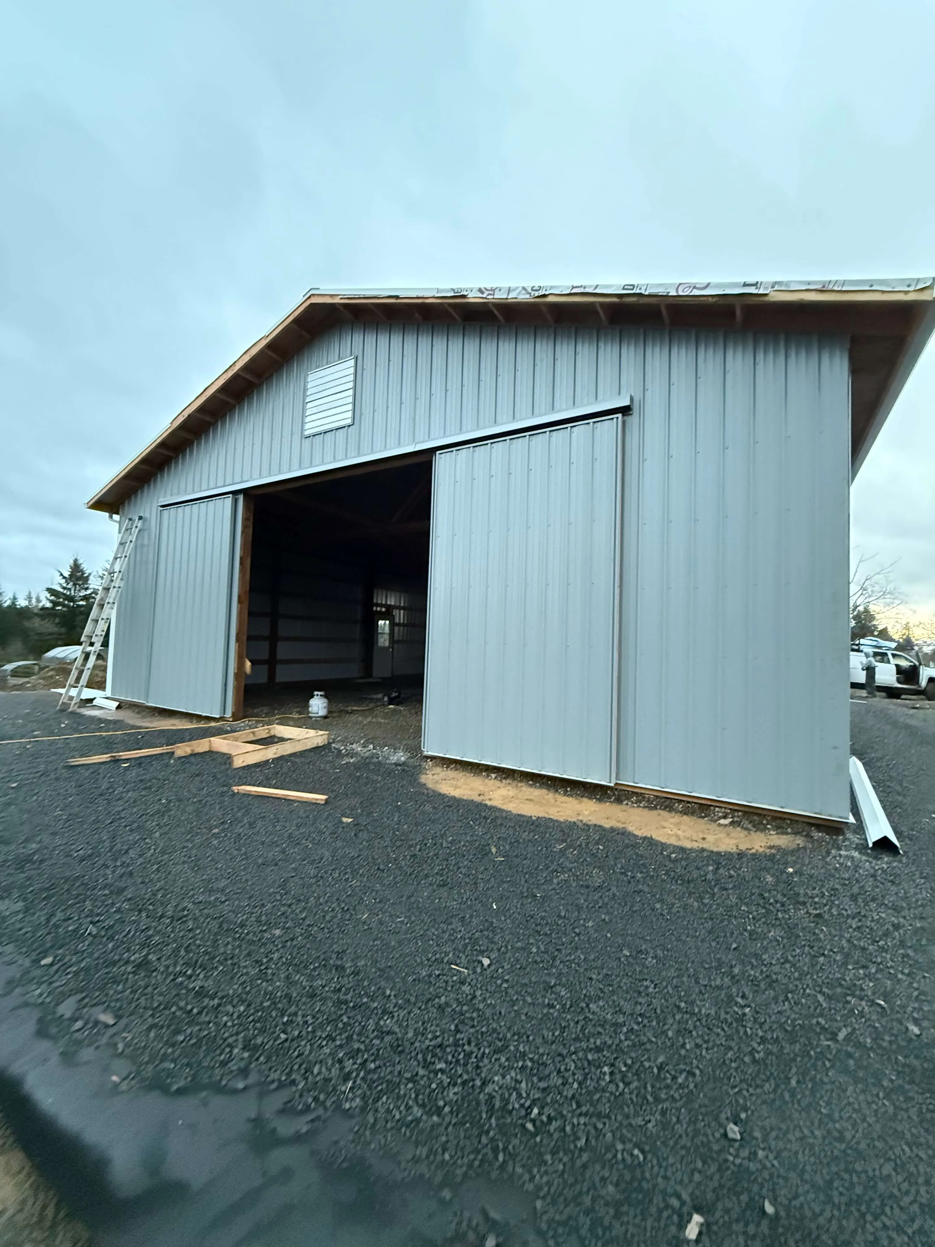 Partially constructed large metal shed with sliding doors open on gravel ground under cloudy sky.