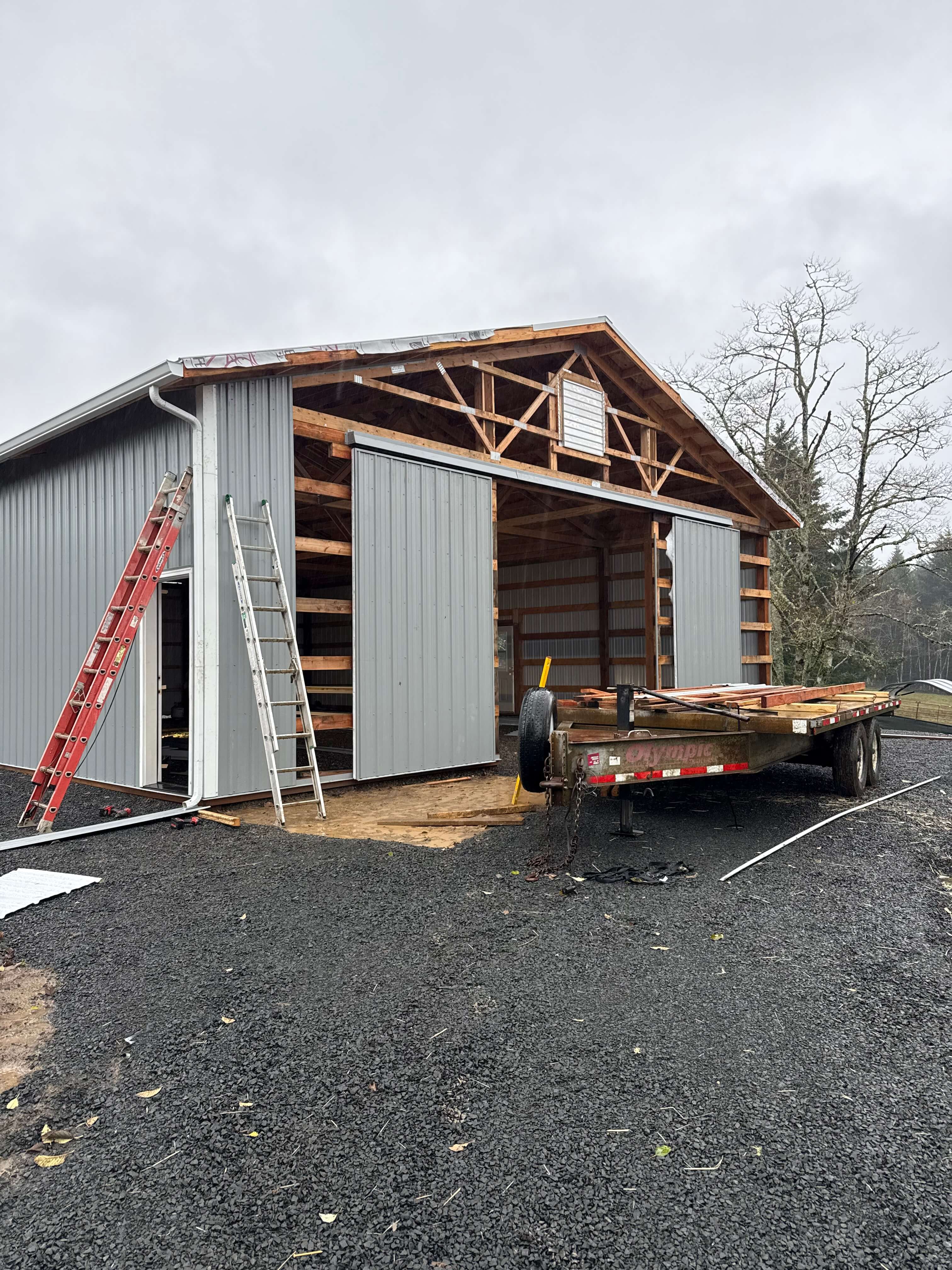 Partially constructed gray metal building with sliding doors open and a trailer loaded with wood in front.