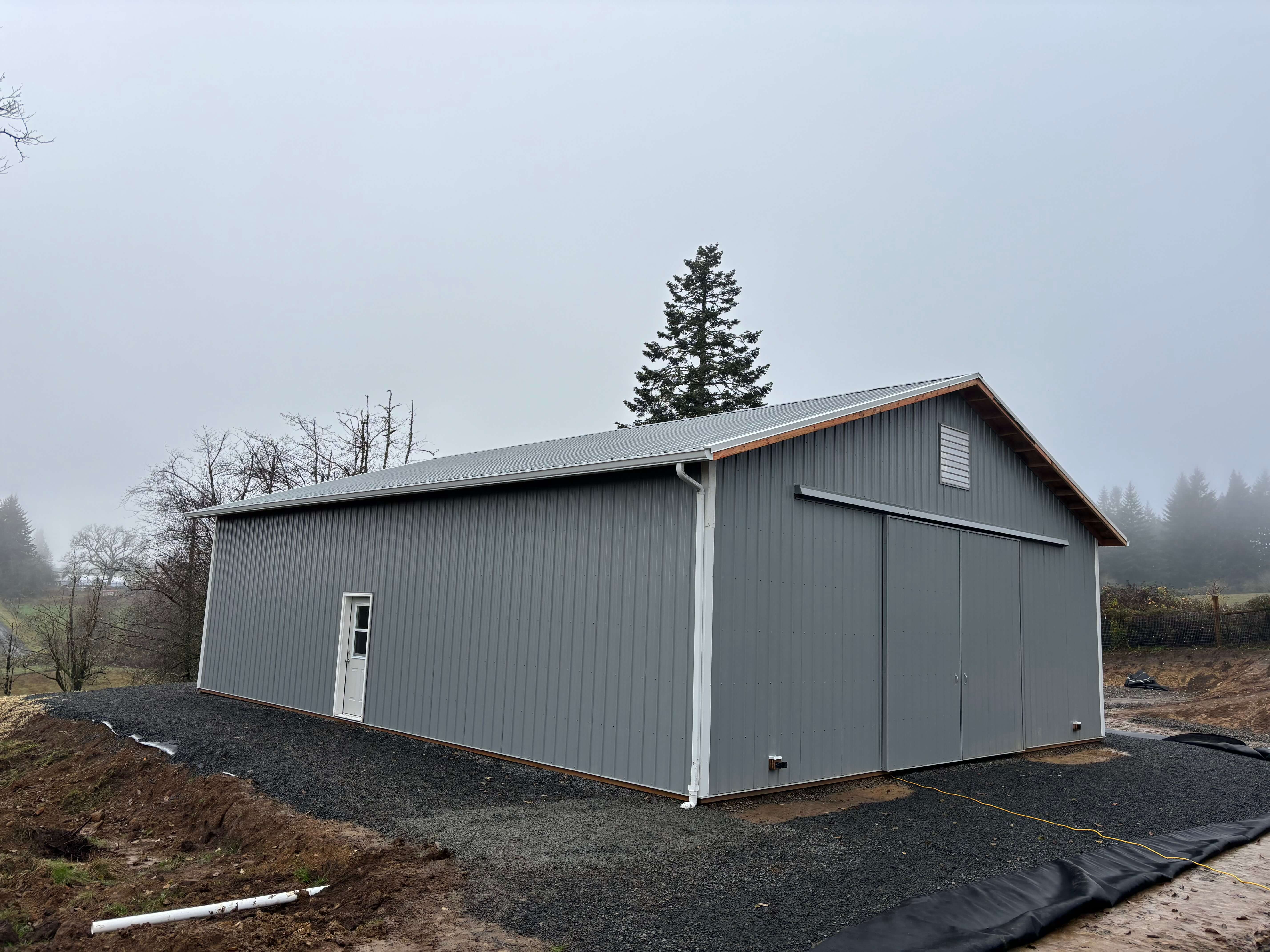 Large gray metal barn with sliding doors and a small entry door on a foggy day.