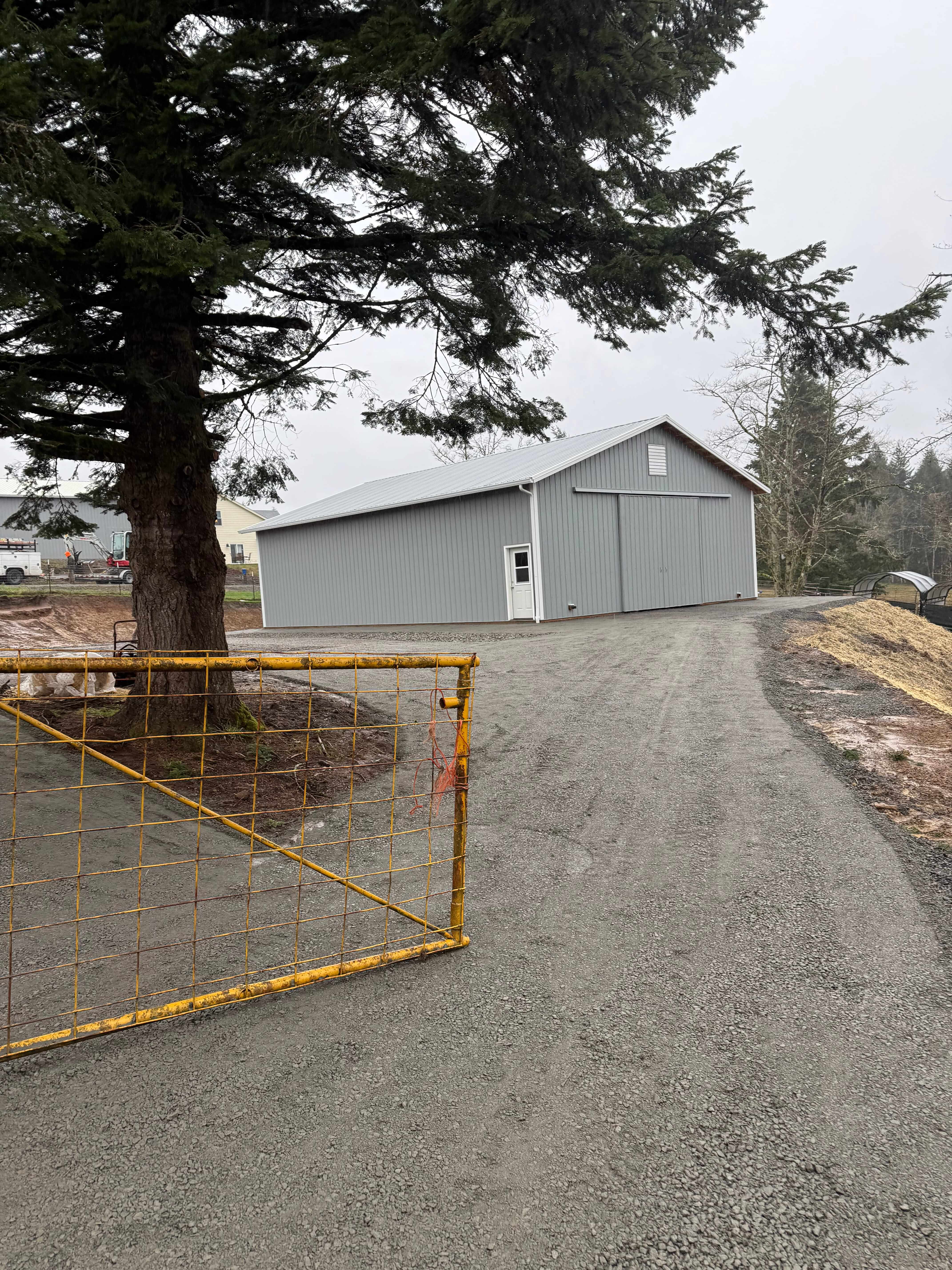 Large gray metal barn with a white door at the end of a gravel driveway near a large tree and yellow metal gate.