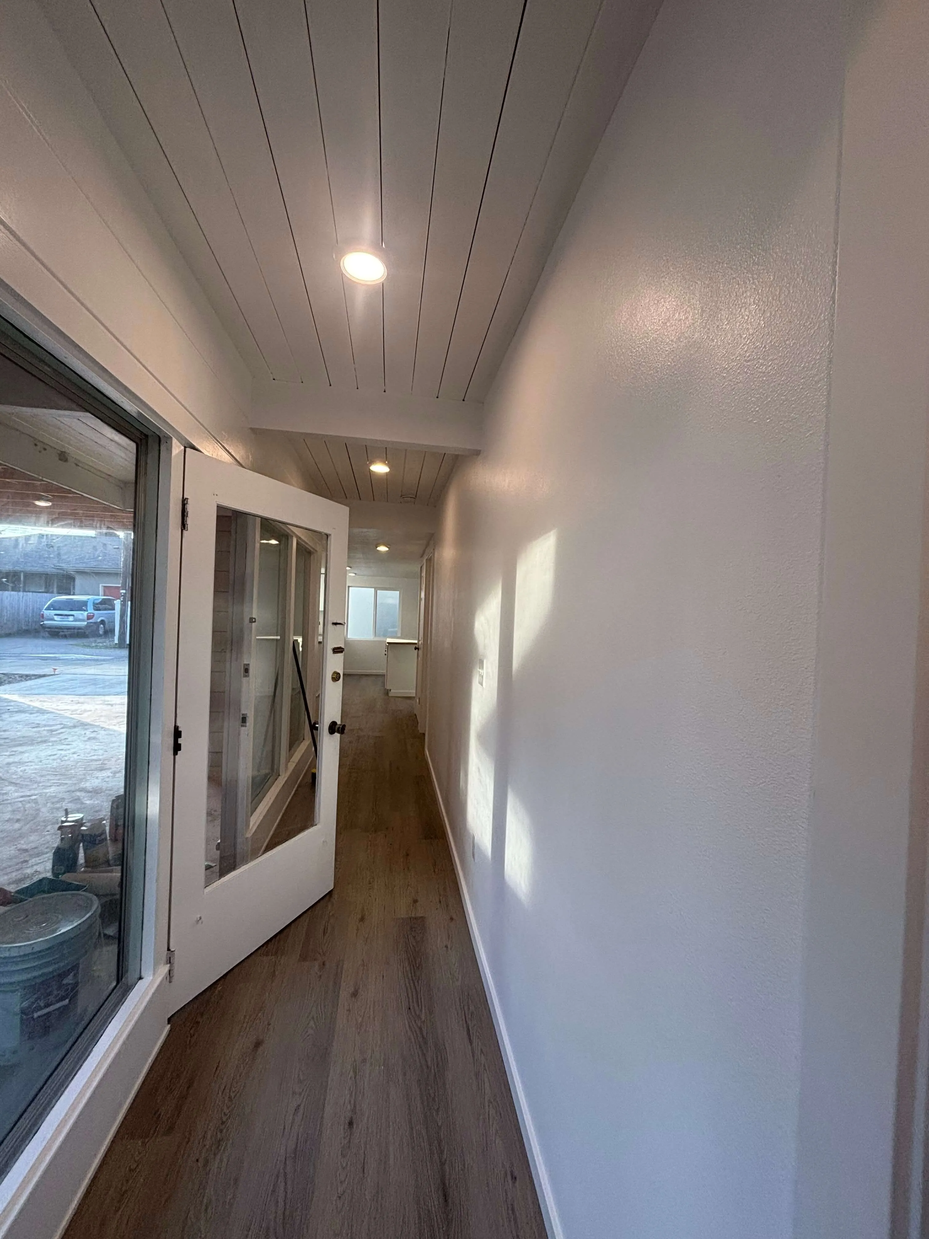 Narrow hallway with wood floor, white walls, recessed ceiling lights, and an open door with large glass panes.