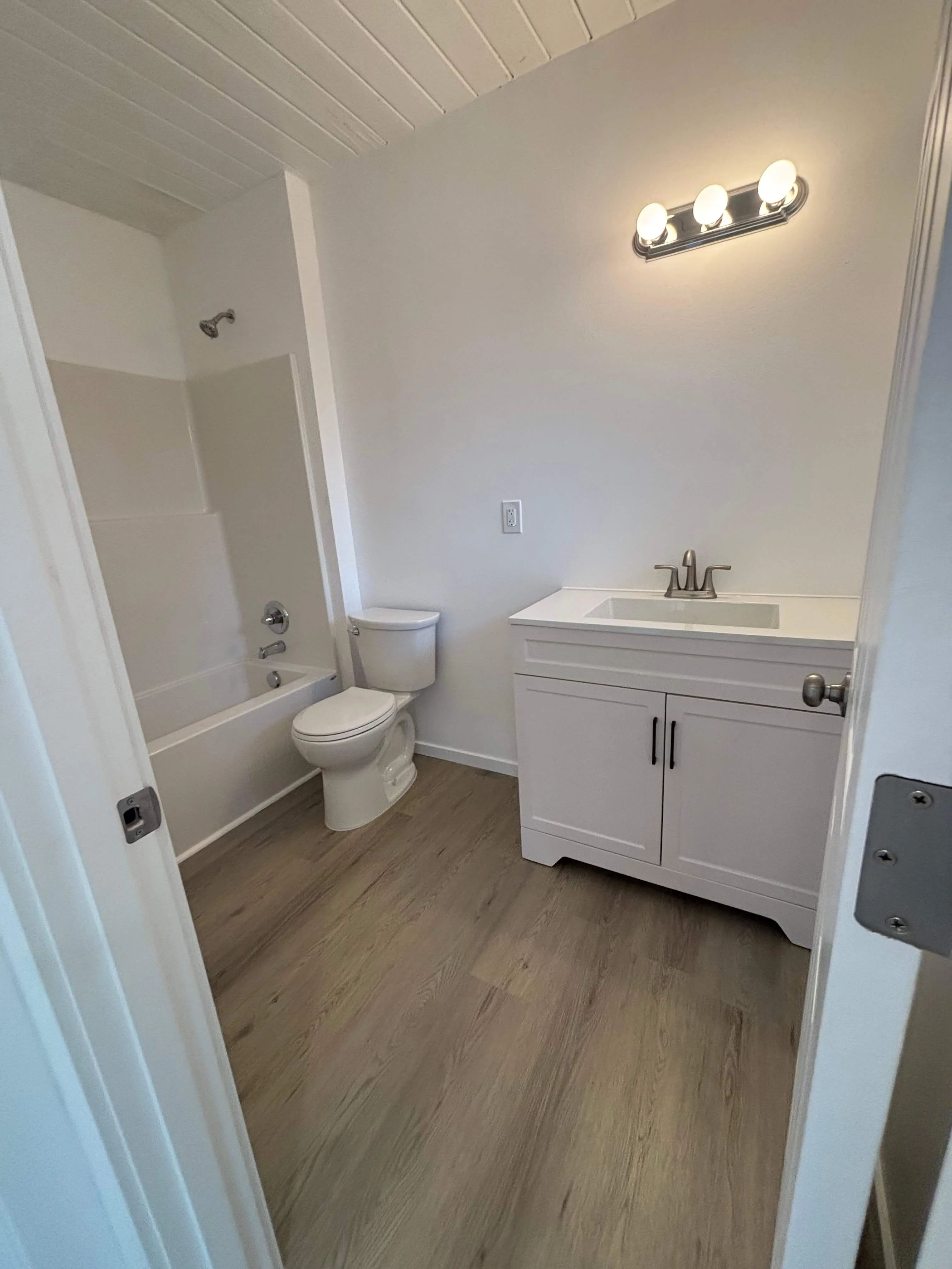 Small bathroom with wood-style flooring, white bathtub, toilet, and vanity with sink and light fixture above.