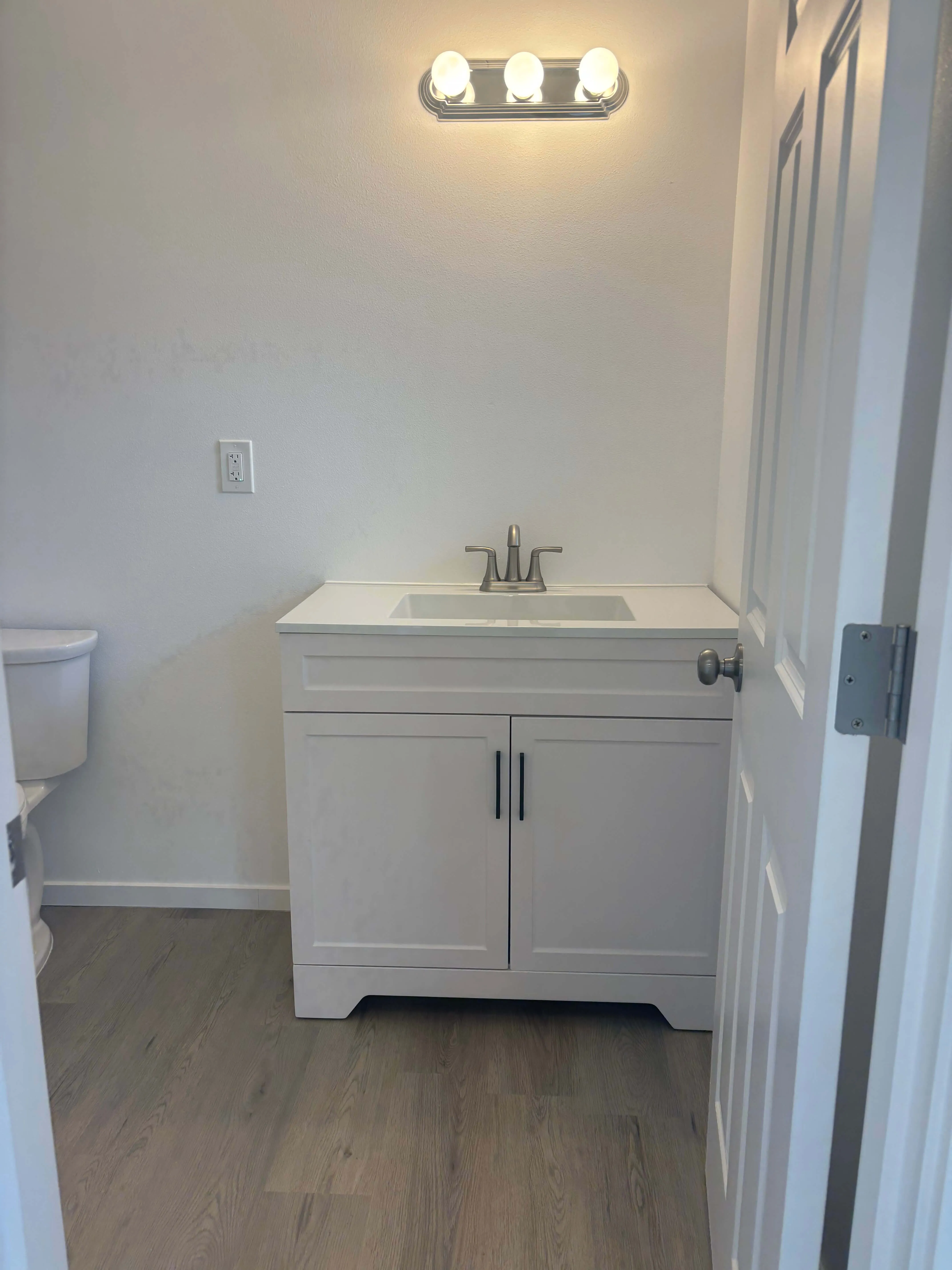 Small bathroom with white vanity, silver faucet, three-bulb light fixture, and partial view of toilet.