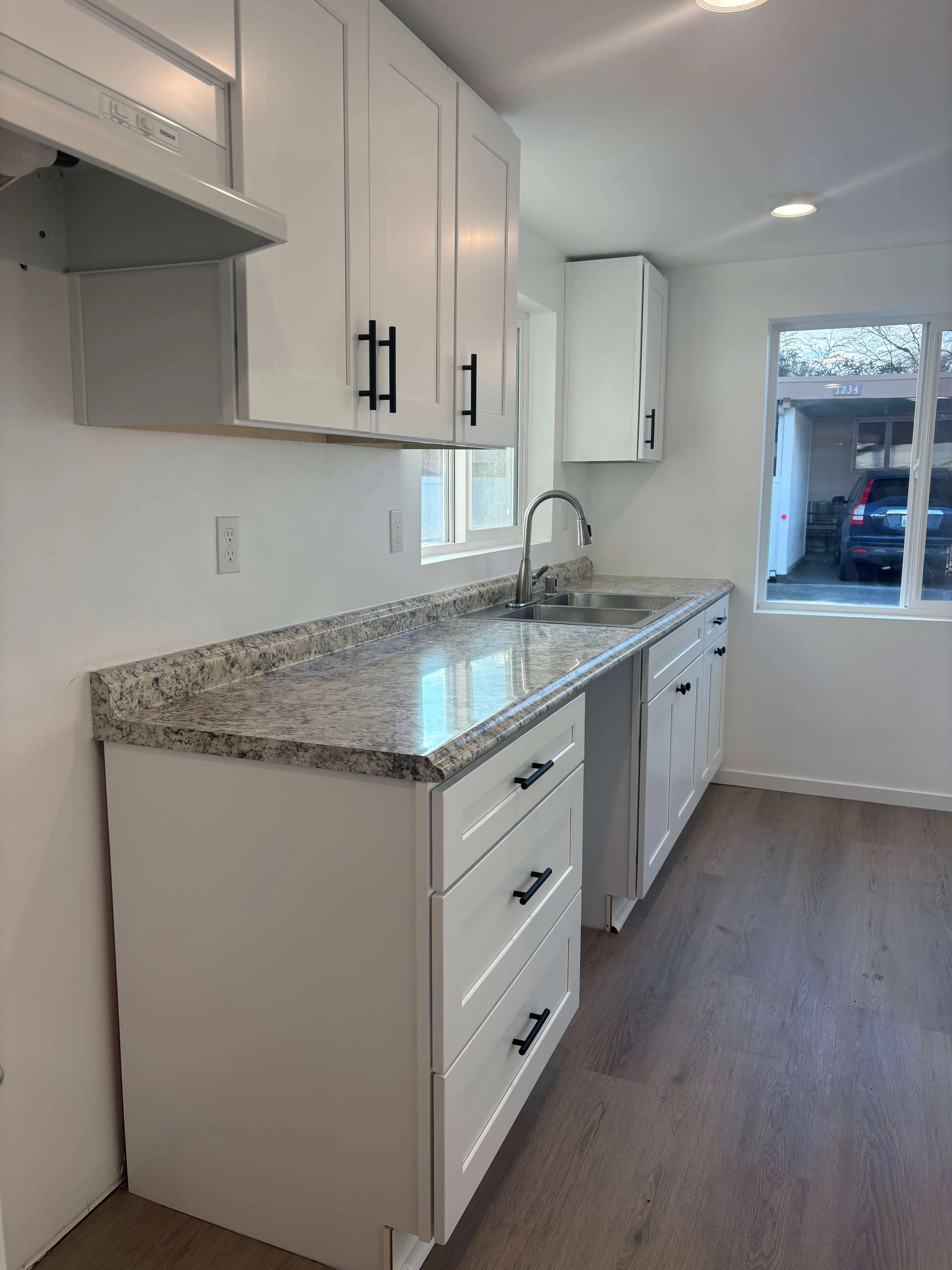 Modern kitchen with white cabinets, black handles, granite countertop, stainless steel sink, and window view of driveway.