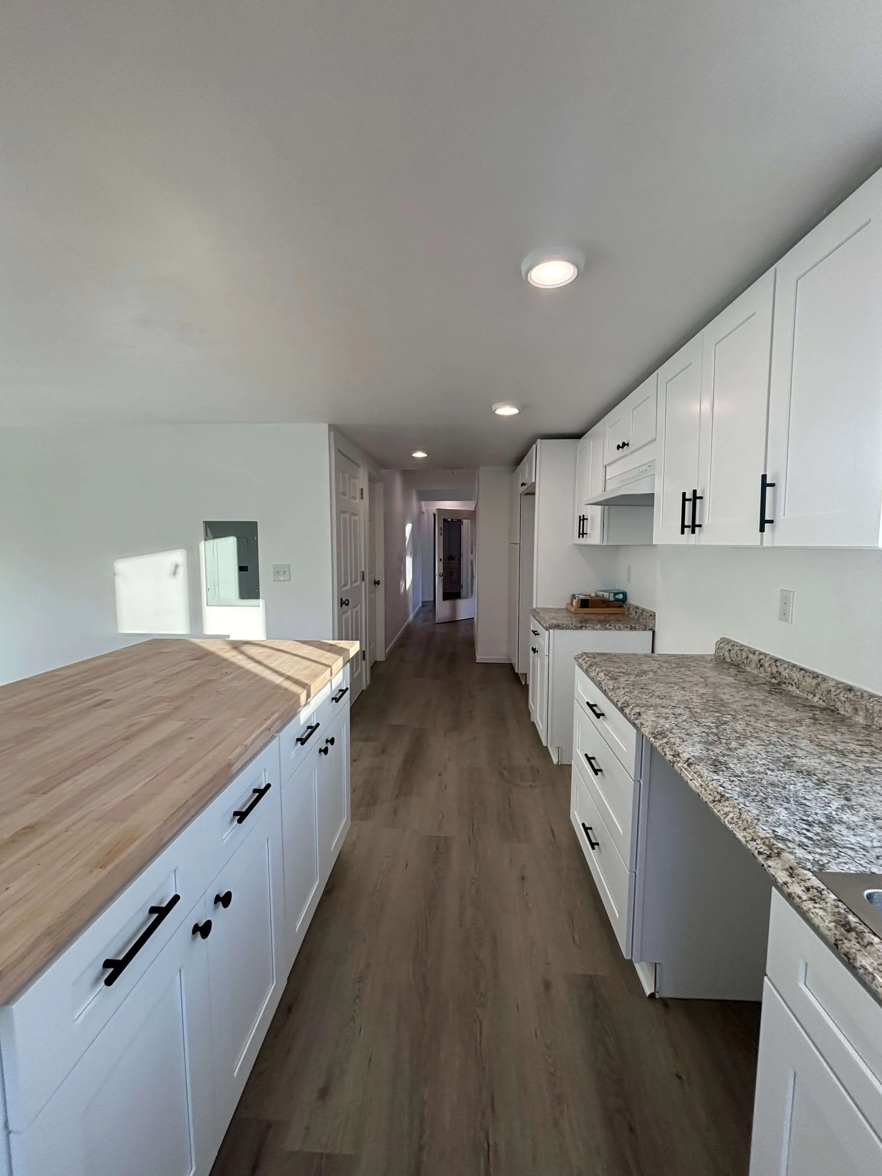 Narrow modern kitchen with white cabinets, wood and granite countertops, and wood flooring.