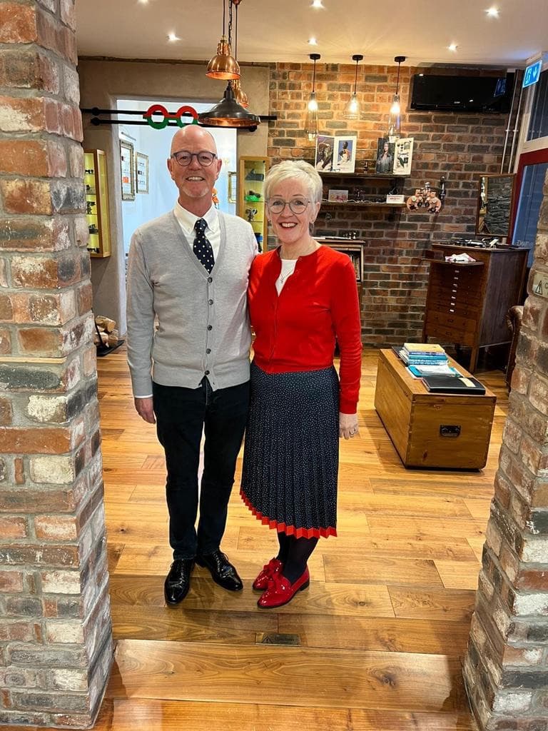 Man and woman standing indoors between brick pillars, smiling toward the camera.
