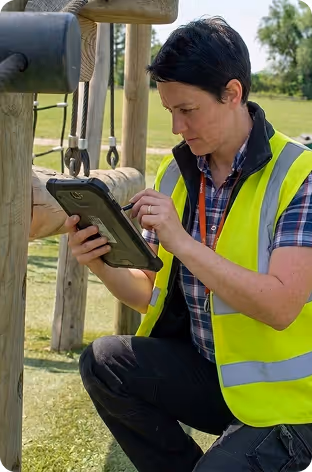 Person in high-visibility vest using tablet near playground wooden climbing frames.