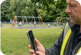 Man in safety vest holding smartphone outdoors with playground and swings in background.