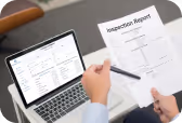 Person holding inspection report form over desk with laptop displaying a spreadsheet.