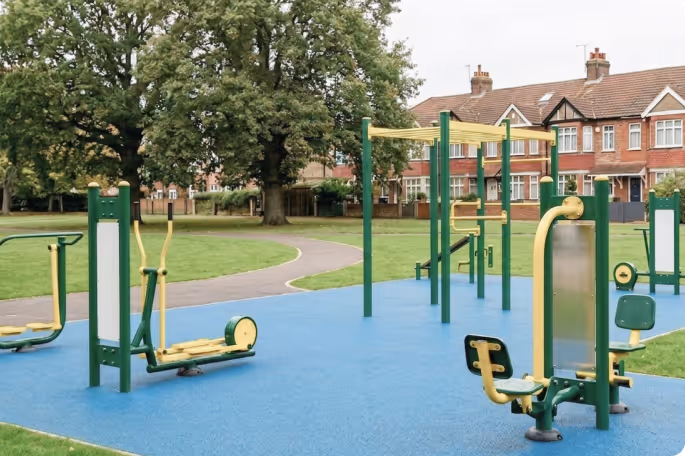 Outdoor fitness equipment on blue flooring in a park with grass, trees, and houses in the background.