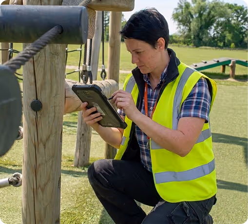 Person in high-visibility vest using a tablet while inspecting a wooden playground structure.