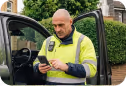 A man in a yellow high-visibility jacket standing by a black vehicle while checking his phone.