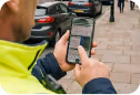 Person using a smartphone on a city sidewalk with parked cars and pedestrians nearby.