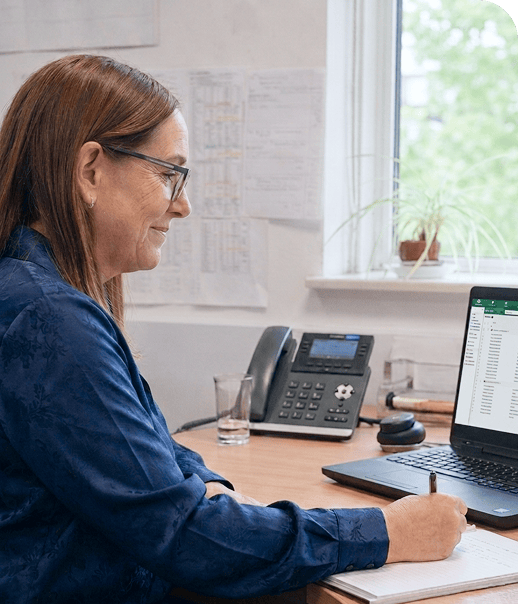 Woman in glasses sitting at desk writing notes while looking at a laptop screen with spreadsheet data.