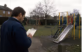 Man writing on clipboard at playground with slide and climbing frame on a cloudy day.