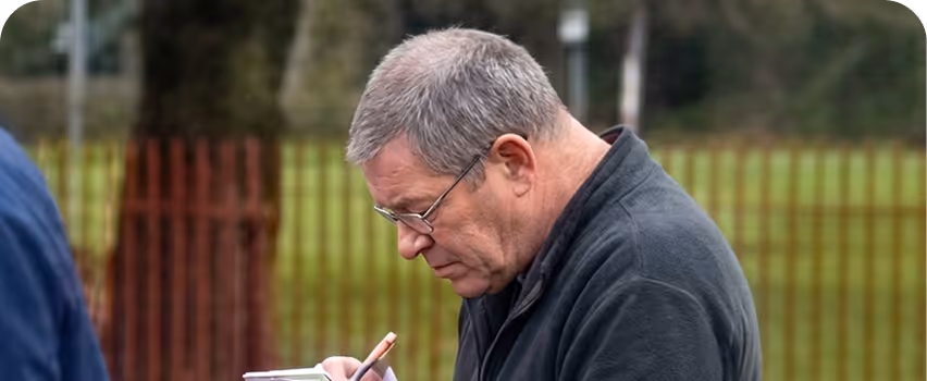 Older man with glasses and grey hair writing on a clipboard outdoors near a fence.