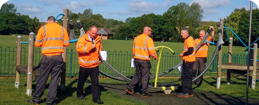 Five men in high-visibility jackets inspecting outdoor playground equipment on a grassy area.